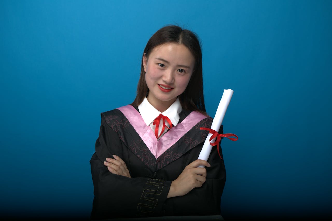Confident graduate in cap and gown with diploma, blue backdrop.